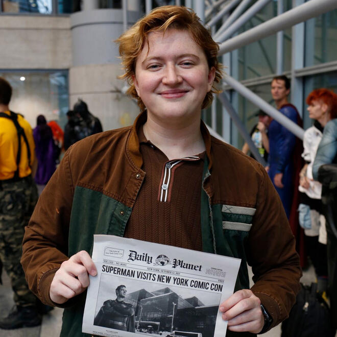 Brie, dressed as Jimmy Olsen from Superman (2025), holding a newspaper and looking into a camera at New York Comic Con.