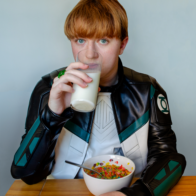 Brie, dressed as Guy Gardner from Superman (2025), sitting at a table drinking from a glass of milk